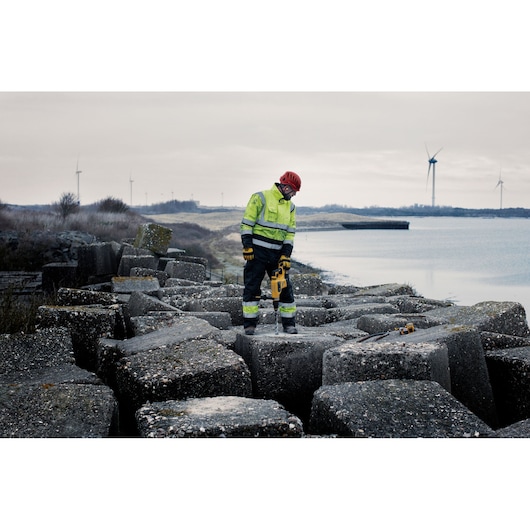 A construction worker wearing a high-visibility jacket and red helmet operates a yellow DEWALT tool on large concrete blocks by the water, with wind turbines visible in the background.