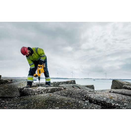 A construction worker wearing a red helmet and green high-visibility jacket is using a yellow DEWALT rotary hammer drill (DCH481N) on large concrete blocks outdoors. Wind turbines are visible in the background under a cloudy sky.