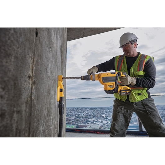 A construction worker wearing safety gear uses a DEWALT cordless rotary hammer drill to bore into a concrete wall on a