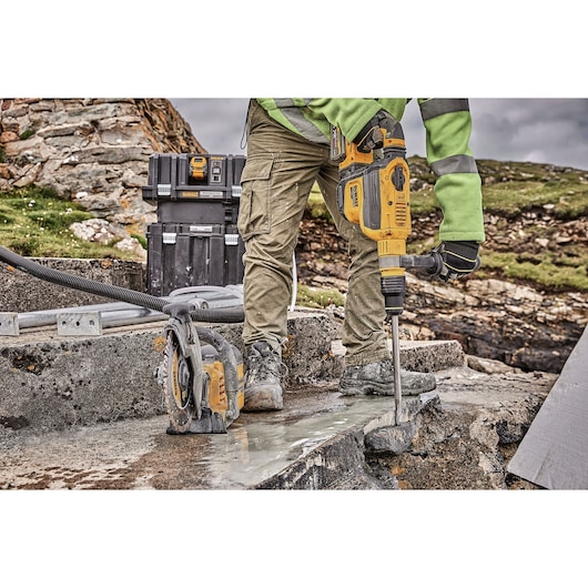 A person wearing work gloves and safety clothing operates a DEWALT heavy-duty rotary hammer drill on a concrete surface