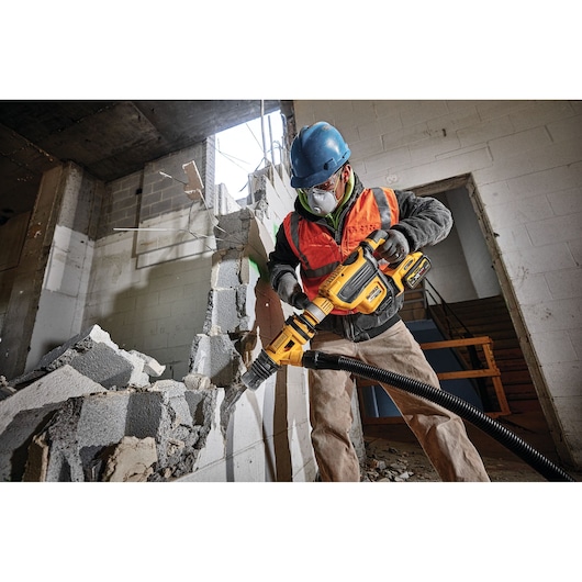 A construction worker wearing a blue hard hat and orange safety vest uses a yellow DEWALT rotary hammer to break down a