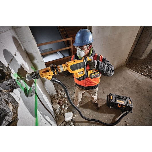 A construction worker wearing a blue hard hat and safety vest uses a DEWALT cordless rotary hammer drill to break through a