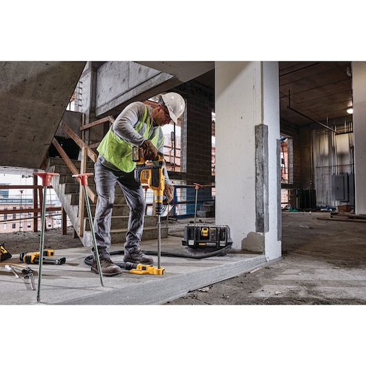 A construction worker using a DEWALT power drill to work on a concrete floor at an indoor construction site. The worker is