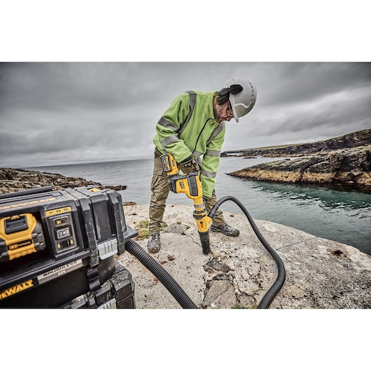 A person wearing safety gear and a green jacket uses a DEWALT power tool to break concrete outdoors near a body of water. A