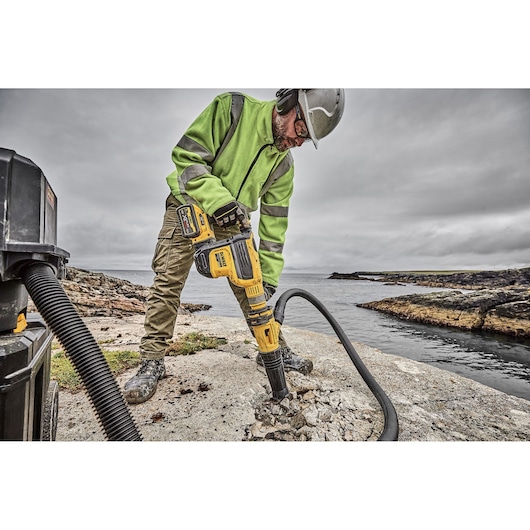 A worker wearing safety gear uses a DEWALT cordless demolition hammer (DCH614X2) to break up concrete outdoors near a water
