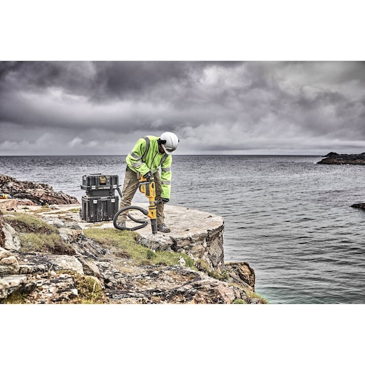 A person wearing safety gear operates a yellow DEWALT rotary hammer drill on a rocky coastal surface with two DEWALT storage