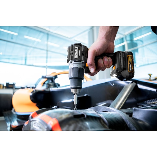 A close-up of a person using a DEWALT cordless drill to fasten or assemble parts on what appears to be a vehicle component, with an orange car visible in the background.