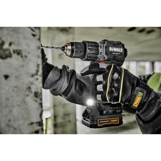 A person wearing protective gloves uses a DEWALT cordless drill to drill into a concrete wall. The drill has a McLaren branded battery pack and an LED light. The scene appears to be in a construction site.