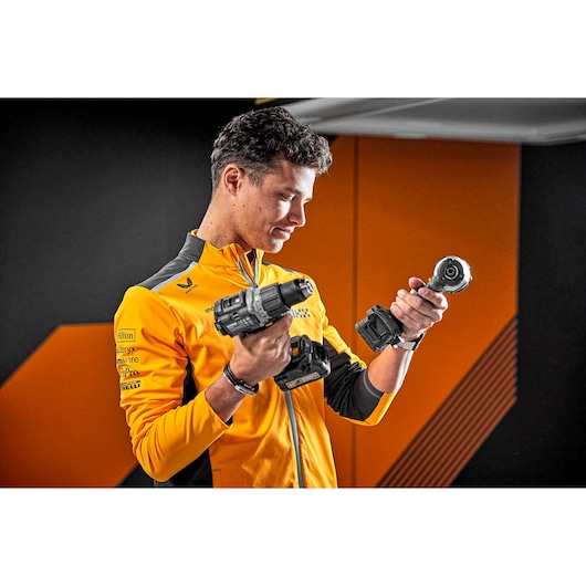 A person wearing a yellow and black jacket holding two DEWALT power tools, standing indoors against a background with black and orange geometric patterns. The person's face is blurred.
