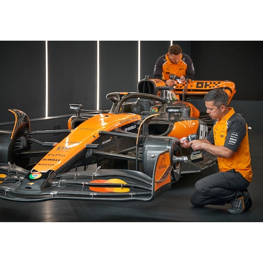 Two people wearing orange and black uniforms are working on an orange Formula 1 race car in a garage. One person is kneeling at the front of the car using a DEWALT power tool near the front wing, while the other is standing and working on the rear of the car.