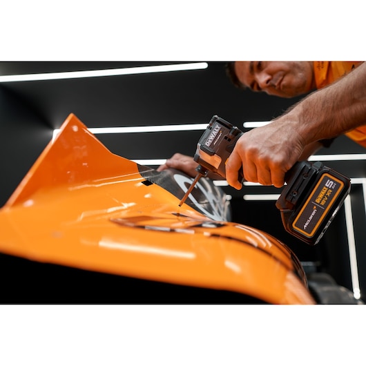 Close-up of a person using a DEWALT cordless impact driver to drill into an orange metal surface, with modern lighting in the background.