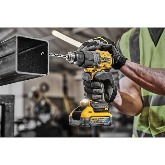 A person wearing gloves and a high-visibility vest is using a yellow DEWALT cordless drill with POWERSTACK battery to drill