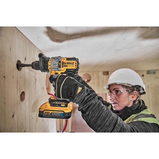 A person wearing a hard hat and gloves uses a DEWALT cordless drill to drill a hole in a wooden wall. The DEWALT Power Stack 18V battery is attached to the drill. The person's face is blurred for privacy.