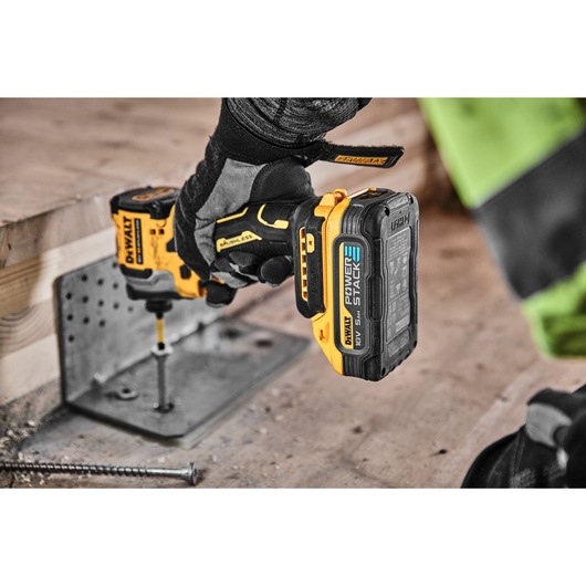 A person wearing protective gloves uses a yellow and black DEWALT cordless power drill with a POWERSTACK battery to drive a screw into a metal bracket attached to a wooden beam at a construction site.