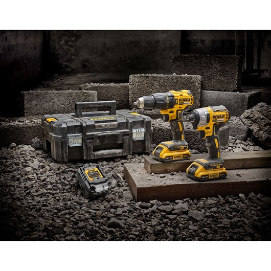 Two DEWALT cordless power tools, carrying case, and charger on a rough, rocky surface near concrete blocks.