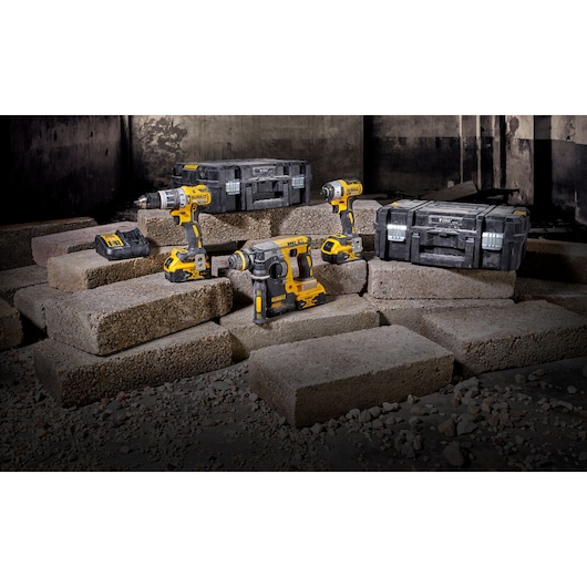 A set of DEWALT cordless power tools is displayed on concrete blocks. The set includes a cordless drill, an impact driver, a rotary hammer, two sturdy black toolboxes, and a battery charger. The tools feature a yellow and black color scheme.
