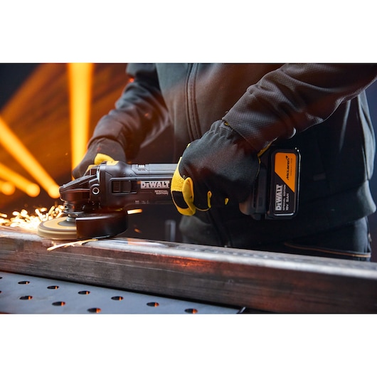 A person wearing gloves is using a DEWALT cordless angle grinder to grind a metal bar, with visible sparks flying from the