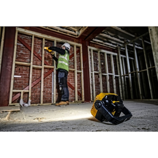 A DEWALT DCL077 work light is placed on the floor of a construction site, illuminating the area while a person wearing