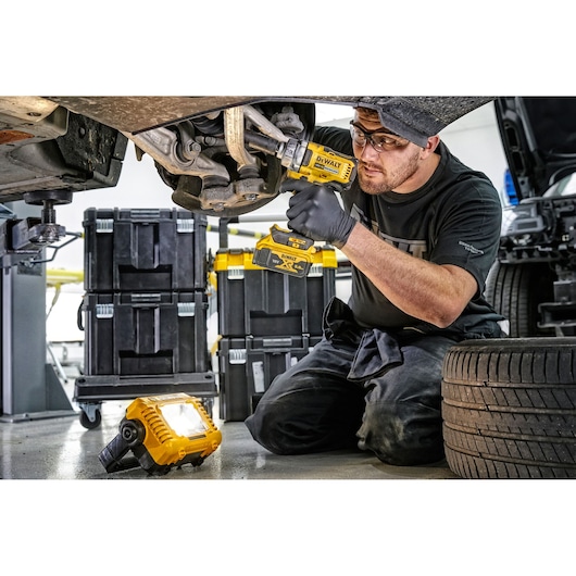 A person wearing black gloves is using a DEWALT cordless power tool to work underneath a vehicle in a garage. A DEWALT