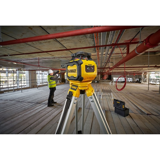 A DEWALT rotary laser level (model DCLE05211R) mounted on a tripod is positioned in the center of a construction site with