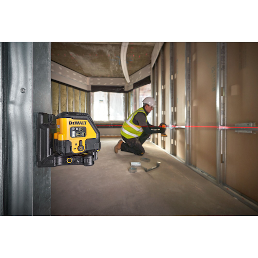 A DEWALT laser level is mounted on a metal frame inside a building under construction. In the background, a person wearing a