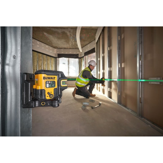 A DEWALT laser level device mounted on a metal frame projecting a green laser line across a construction site interior. A
