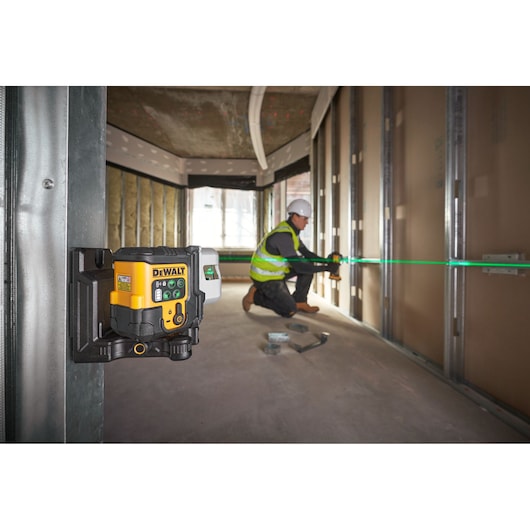 A DEWALT laser level device (SKU: DCLE14301GB) mounted on a metal frame inside a building under construction. The laser projects a green line across the room, while a person in safety gear works with tools along the line.