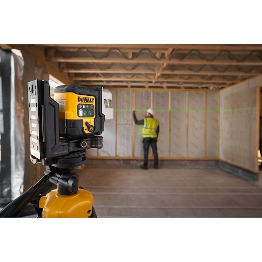 A DEWALT laser level device mounted on a tripod is in use at a construction site, projecting a green horizontal laser line. A person wearing a safety vest and helmet is working near a wall in the background.