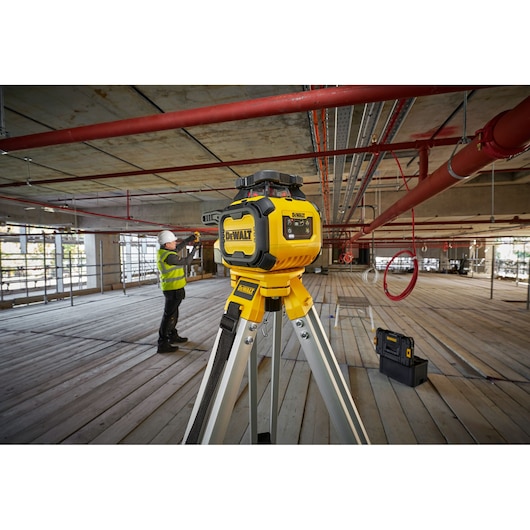 A DEWALT laser level device mounted on a tripod is positioned in the foreground of a construction site with exposed pipes and scaffolding. In the background, a worker wearing a hard hat and a yellow safety vest is taking measurements. A DEWALT case is placed on the floor nearby.