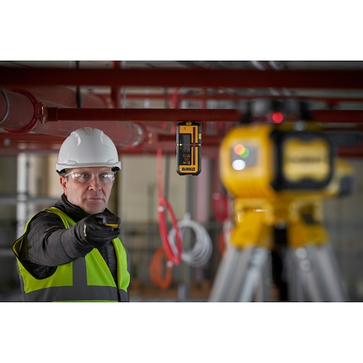 A person wearing a white hard hat and a high-visibility vest is using a DEWALT laser receiver in an industrial setting. A DEWALT laser level device is mounted on a tripod in the foreground, and the surroundings include red pipes and cables.