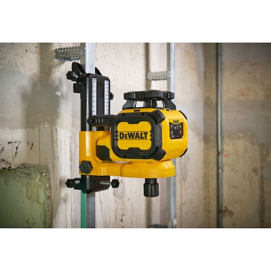 A DEWALT yellow and black rotary laser level device mounted on a metal bracket, attached to a vertical metal stand, positioned against a concrete wall in a construction setting.