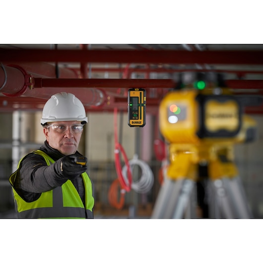 A person wearing a safety vest and helmet is using a DEWALT laser detector (model DCLE15221G) on a construction site. The laser detector is attached to a metal pipe, and a yellow DEWALT laser level is visible on a tripod in the foreground.