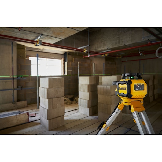 A DEWALT laser level device is positioned on a tripod in a construction site, projecting a green horizontal line across the unfinished room with concrete blocks and exposed pipes.