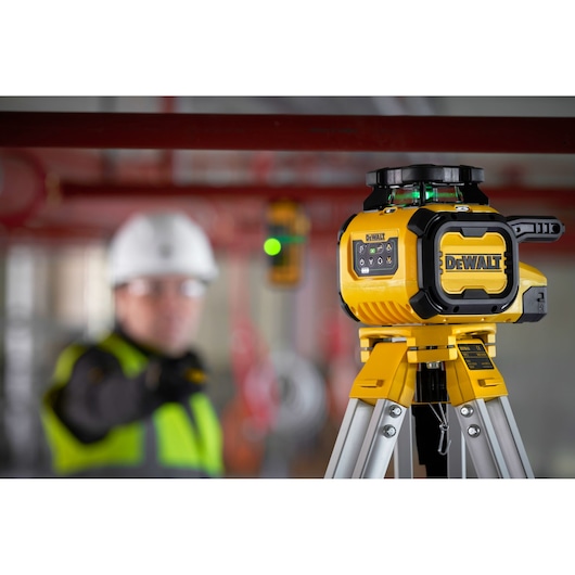 A DEWALT DCLE15221G laser level device mounted on a tripod is shown in the foreground of a construction site. In the background, a person wearing a hard hat and high-visibility vest is standing, but their face is blurred for privacy.
