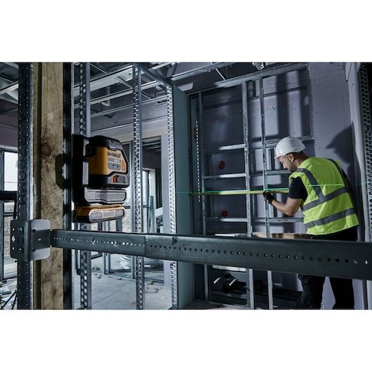 A DEWALT laser level is mounted on a wooden post in a construction site, projecting a green laser line towards a worker
