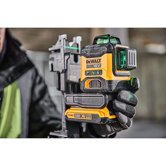A person wearing black gloves is holding a DEWALT 18V laser level tool with a visible green laser. The device has a yellow and black design, with control buttons on the front and an 18V XR Li-Ion battery pack attached at the bottom.