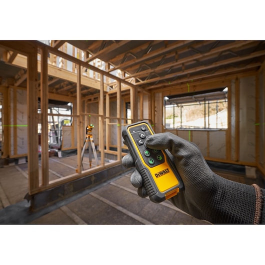 A gloved hand holds a DEWALT remote control in the foreground, with a DEWALT laser level on a tripod projecting green lines