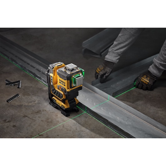 A DEWALT laser level tool with green laser lines is shown on a concrete floor, being used to align metal beams. A person