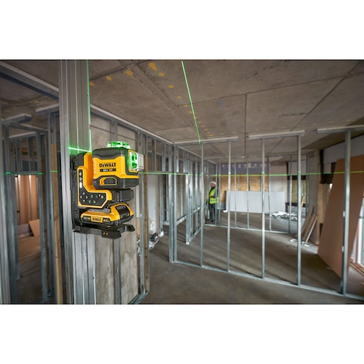 A DEWALT green line laser tool mounted on a metal frame inside a building under construction. The laser projects green lines