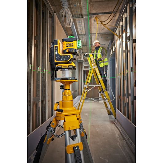 A DEWALT laser level (model DCLE34035D1) mounted on a tripod in an indoor construction site, projecting green laser lines