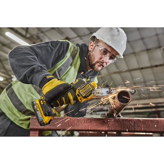 A person wearing safety gear uses a DEWALT cordless power tool to cut through a metal pipe, with sparks flying from the cutting action in an industrial setting.