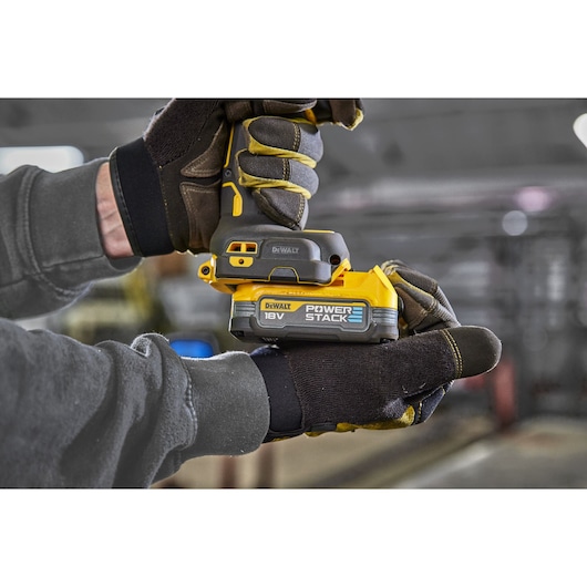 A close-up image showing gloved hands holding a DEWALT 18V POWERSTACK battery attached to a cordless tool, in an indoor workshop setting.
