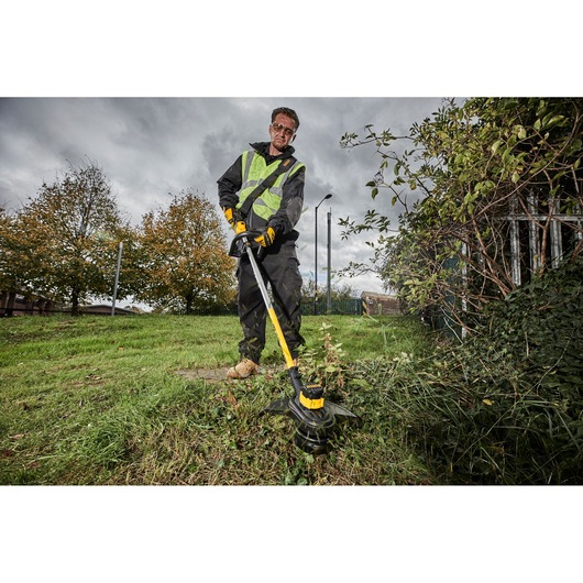 Person using a DEWALT grass trimmer outdoors, wearing safety gear and trimming grass near bushes.
