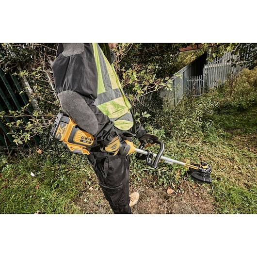 Person using a DEWALT cordless grass trimmer outdoors, cutting tall grass near a fence.