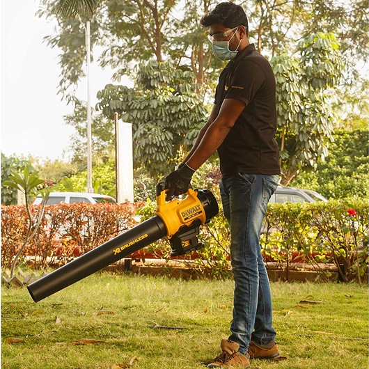 A person outdoors holding a yellow and black DEWALT DCM562P1 cordless blower, wearing gloves and standing on grass with trees and shrubs in the background.