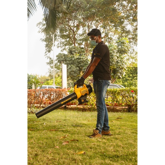 A person using a DEWALT DCM562P1 cordless blower outdoors on a lawn, surrounded by trees and bushes. The person's face is blurred for privacy.