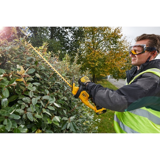 A person wearing safety gear and gloves is using a DEWALT DCM563PB cordless hedge trimmer to cut a dense bush outdoors. The person's face is blurred for privacy.