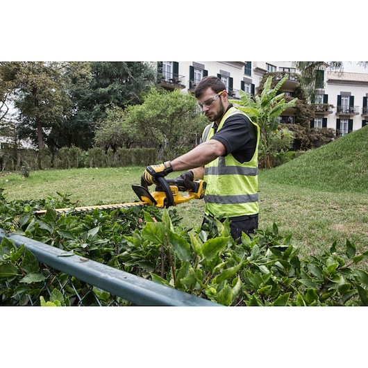 A person wearing a fluorescent safety vest and gloves using a yellow DEWALT cordless hedge trimmer to trim bushes in a garden area, with residential buildings in the background.