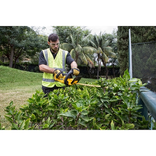 A person wearing a high-visibility vest is using a DEWALT DCM563PB cordless hedge trimmer to trim bushes outdoors. The person's face is blurred for privacy and they are wearing protective gloves.