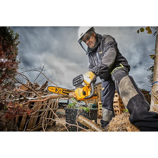 A person wearing safety gear uses a DEWALT DCM565N cordless brushless chainsaw to cut tree branches outdoors. The scene features cloudy skies, wooden pallets, and protective equipment.